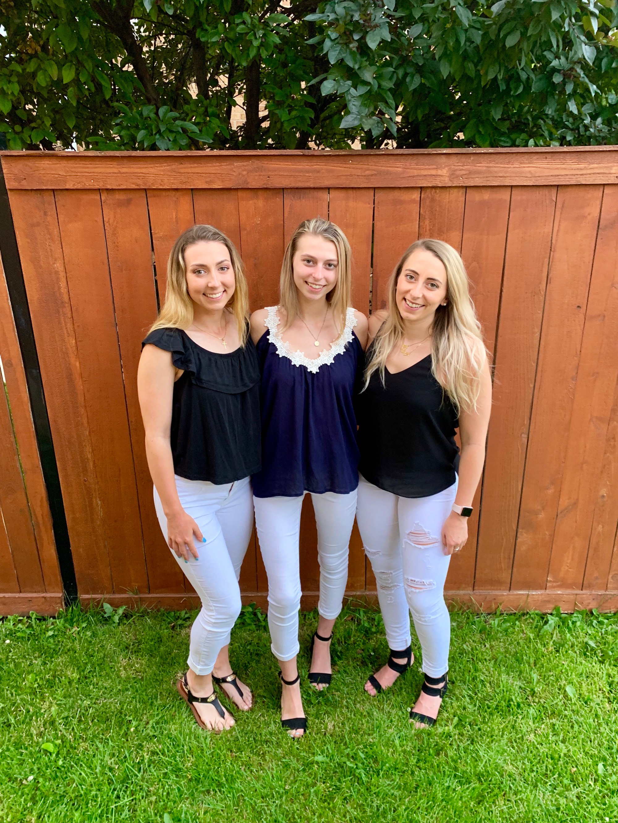 Three sisters all wearing black tops and jeans standing together in from of a wood fence